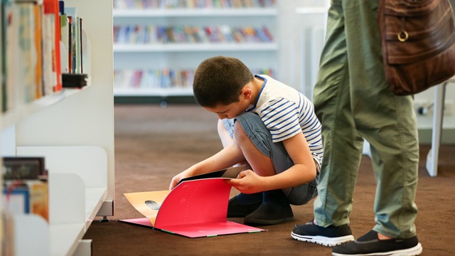 Young boy flicks through a book while kneeling on the floor in a library.