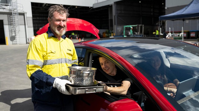 A man in a high-visibility jacket holds a metal container as a person in a red car reaches out to pass it over, outside a large building with an open door.
