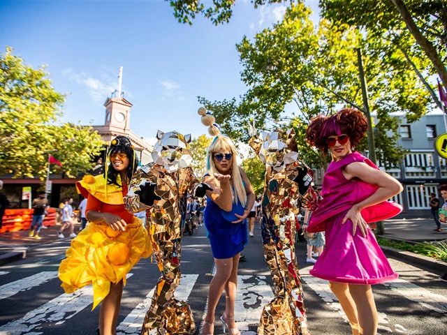 Colourful drag queens and human sized tigers stand on a pedestrian crossing on a sunny day and pose looking at the camera.