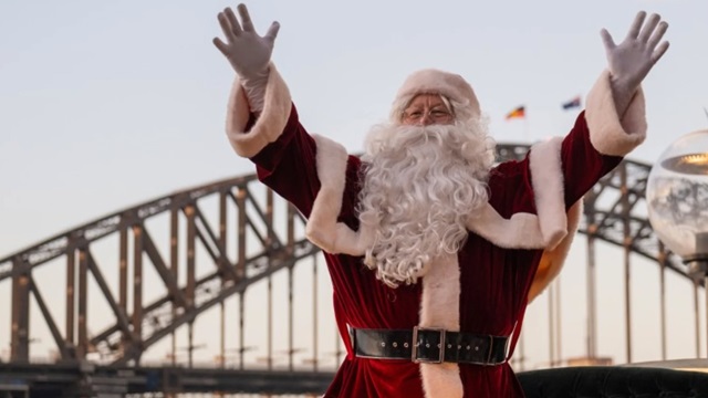 A person in a Santa Claus costume stands outdoors with arms raised, with a large bridge and two flags visible in the background.