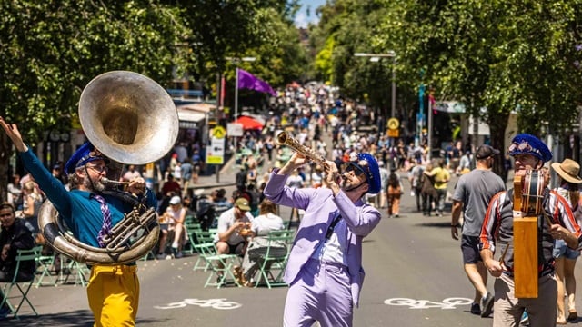 Three musicians perform with brass and percussion instruments on a sunny street closed to traffic, as a crowd walks and sits in the background amid trees and outdoor seating.