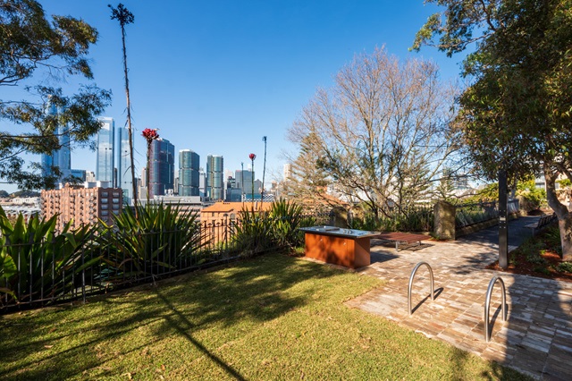 A scenic view of a city skyline from a park with lush green grass. In the foreground, there’s a well-maintained lawn bordered by dense shrubbery and plants. A wooden bench and a metal handrail are visible, suggesting a walking path or viewpoint. The middle ground shows more greenery and trees with autumnal foliage. In the background, modern high-rise buildings stand under a clear blue sky. 