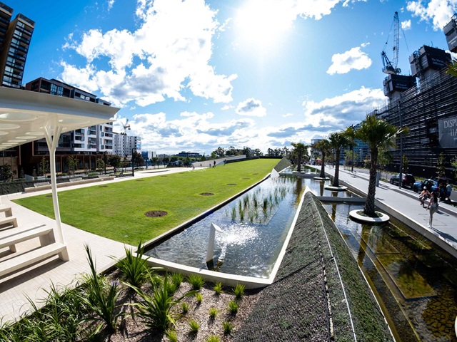 Barbecue area, paving and sloping landscaped surfaces around the lawn. Trees planted in the wetland water feature that runs down the side of the park. Next to it is Zetland Avenue. Apartment buildings around the park in the background.