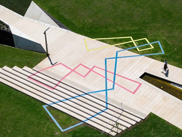 People walking on top of the Stream artwork, embedded in the paving, the artwork is made up of pink, yellow and blue intersecting, asymmetric diamonds that light up at night.