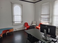 A small room with 2 windows, a boomerang shaped seat with 2 orange single seats, 1 white seat and a desk with a monitor and laptop on top with 2 black seats around the desk
