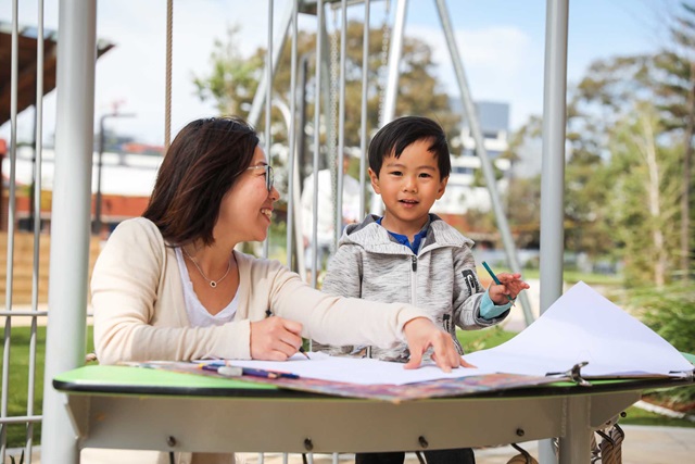 A mother and son at a table with papers doing craft, behind them are steel structures and trees.