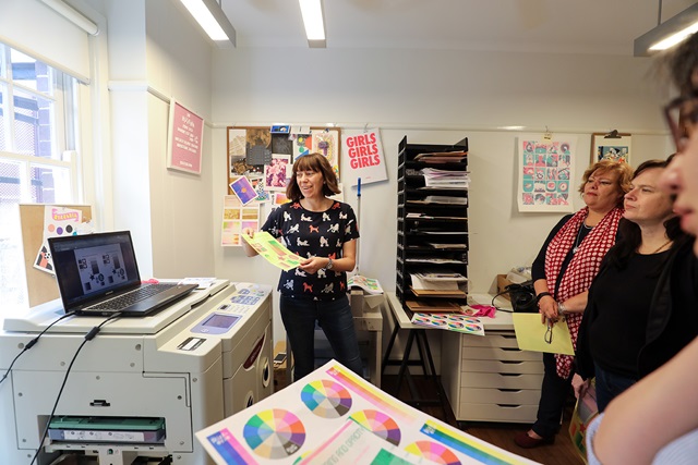 A person stands by a printer, holding printed materials and presenting to a small group in an office decorated with posters and charts.
