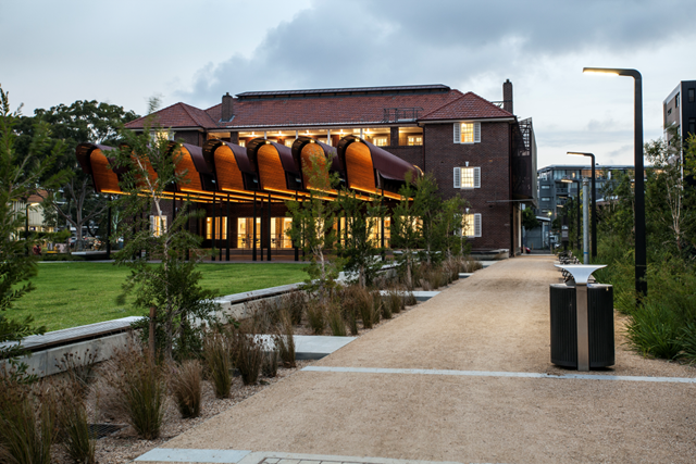 A modern building with a curved wooden canopy and large windows, surrounded by greenery and a gravel path, photographed at dusk.