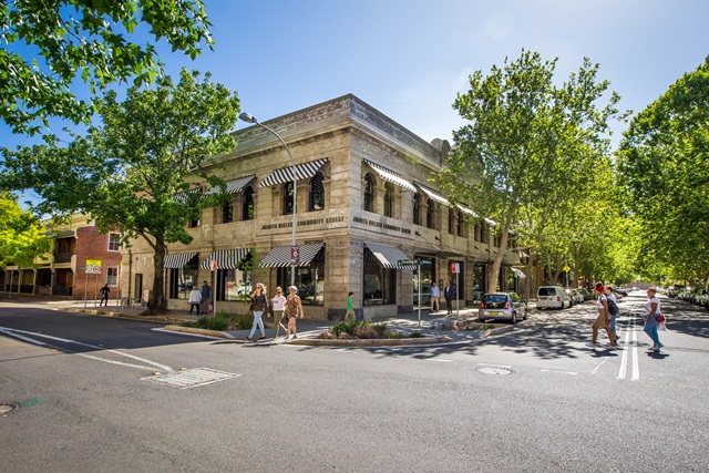 A 2 storey large building on a corner with trees around it and a road in front.