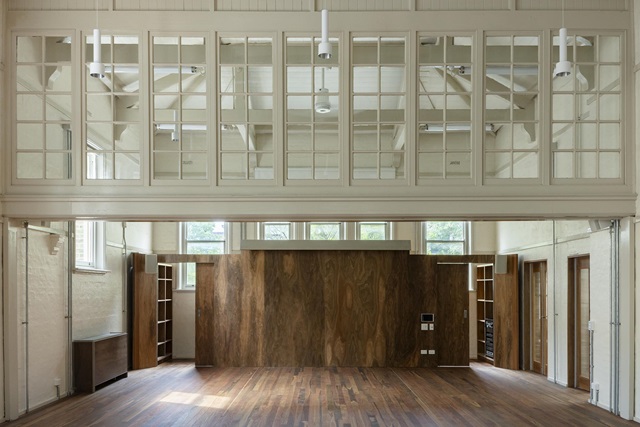 Spacious interior with large wooden floor, high ceilings, and divided glass windows above. Walls feature wood and light tones, with several doorways and natural light from windows.