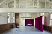 A spacious room with high ceilings features purple curtains, two stacked black chairs, and a small table. Large windows allow natural light to fill the area.