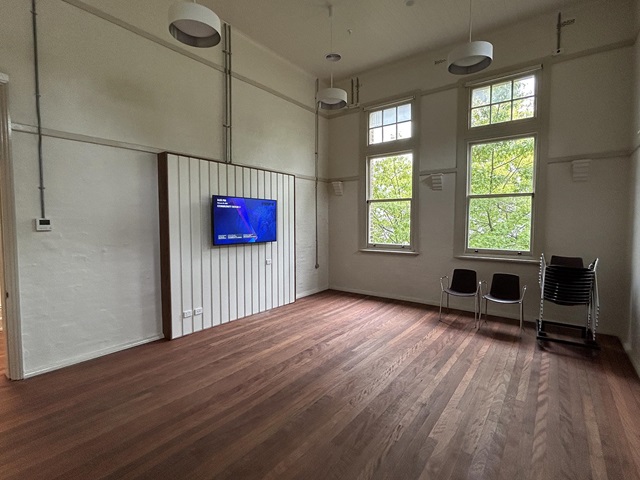 Empty room with wooden floor, wall-mounted TV, two stacked brown chairs, and large windows showing green trees outside