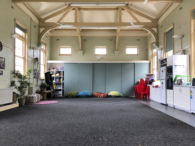 A large open room with high ceilings, carpeted floor and a kitchenette against the wall.