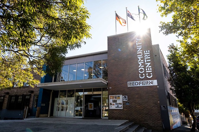 Redfern Community Centre viewed through foliage, with sun peeking over the roof. The 2-story building has a brick front, large windows, and 3 flags on the roof.