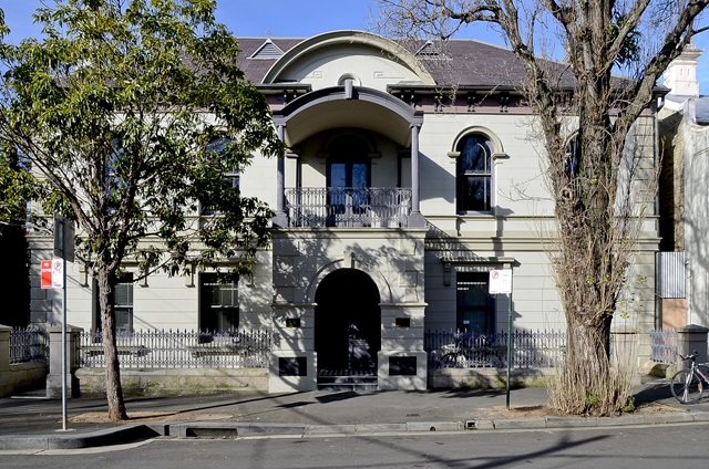 A large 2 storey older building, facing out on to a public path and road with trees in front.