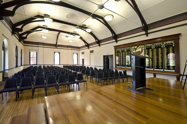 Large open hall with wooden floor boards, a high ceiling and neatly ordered chairs facing a raised podium.