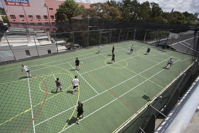 Aerial view over a group of people playing futsal on an outdoor multi-purpose sports court, covered by a protective net.