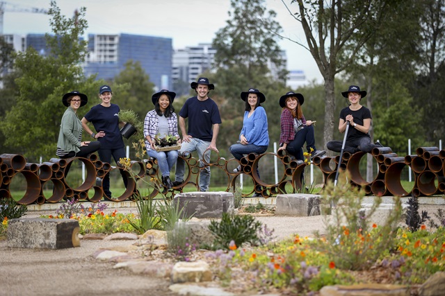 Plants in the foreground, and in the background is a group of 7 adults all wearing hats and holding plants and tools.