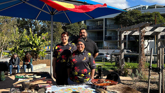 three people wearing Aboriginal colours under a market umbrella flying the Aboriginal flag and behind a market stall displaying Aboriginal artefacts. Plants and building in the background.