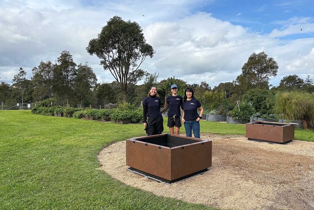 Three people stand together outdoors on grass beside a square raised garden bed, with trees and cloudy sky in the background.