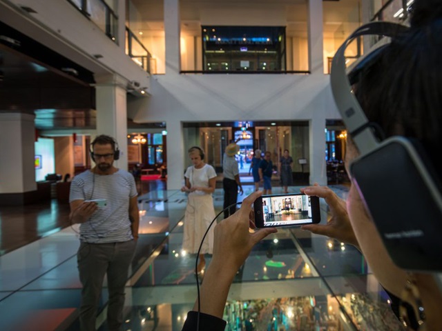 Woman inside Customs House wears headphone connected to her smartphone. It's twilight, the room is lit up. The woman looks at the smartphone screen. Other people standing in the background do the same thing.