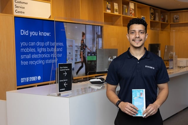 A man in a City of Sydney uniform stands in a customer service centre, holding a leaflet. A sign in the background provides information about electronics recycling.