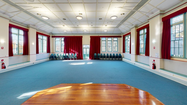 An empty room, with wooden dance floor and blue carpet, long red curtains and mutiple stacks of chairs against the back wall.