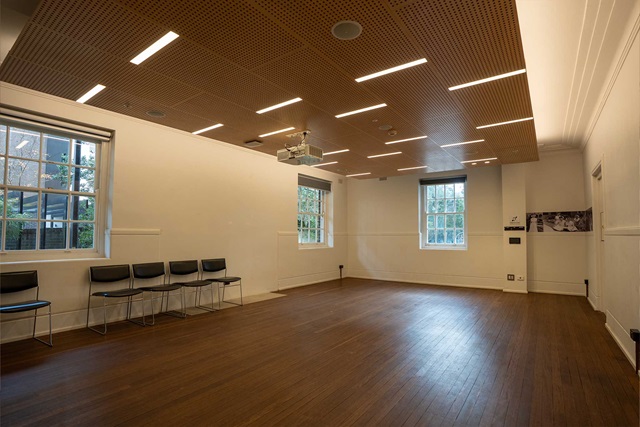 An empty meeting room with wooden floor and 3 windows. 5 chairs are lined against the walls