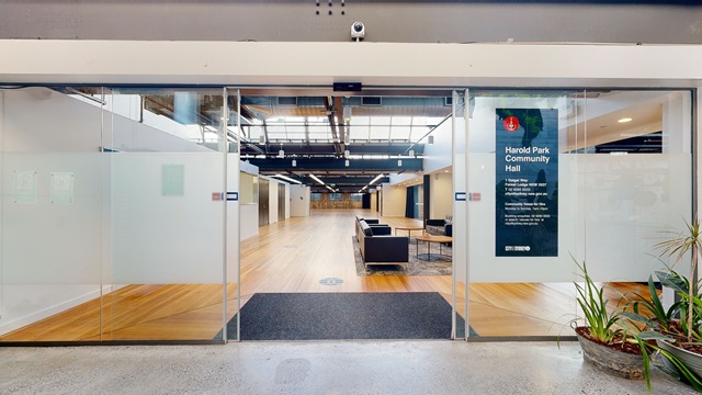 Open glass front door with Harold Park Community Hall sign on right, looking into the space with wooden floor and black lounge