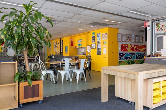 A brightly lit classroom with yellow lockers, white chairs around black tables, educational posters on the wall, a wooden table and storage unit, and a potted plant.