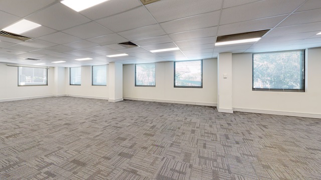 An empty room with large windows and grey carpet.