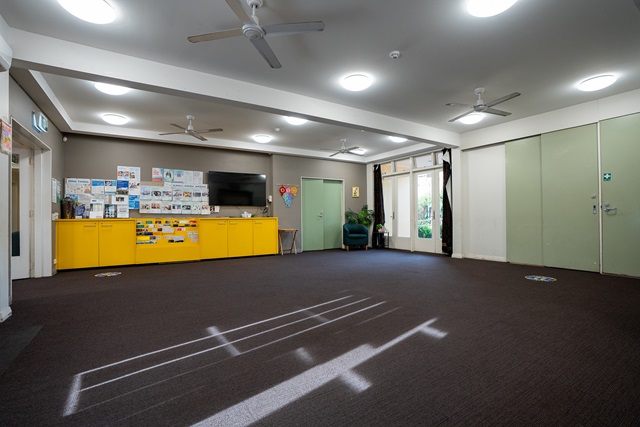 Spacious room with brown carpet, yellow cabinets, wall-mounted TV, bulletin boards, green doors, ceiling fans, and bright overhead lights.