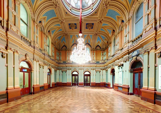 An large ornate room with wooden floor, chandelier, painted walls and features.