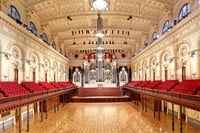 View from mezzanine of a very large heritage hall with stage and round tables dressed for a function.