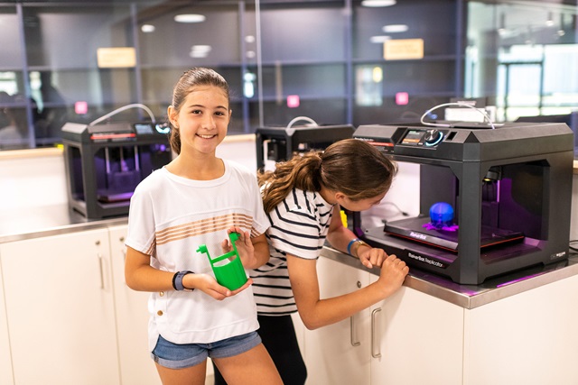 Two girls stand in a makerspace with white cabinets and 3D printers. One girl looks at an item being 3D printed while the other holds a finished item and smiles.