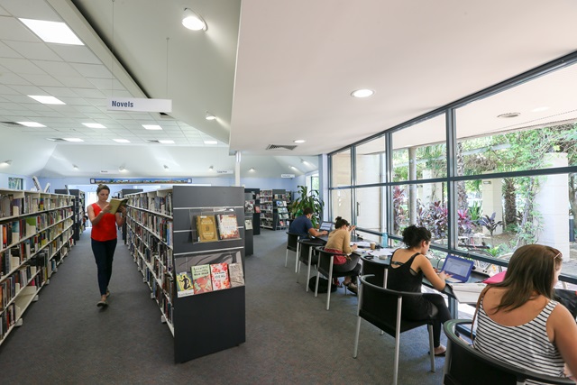 Inside a library there are rows of books, and people sitting at computers near large glass windows on the right.
