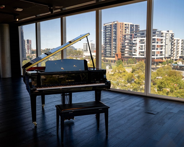 a grand piano on wooden floors with floor to ceiling windows and trees and high rise building in the background