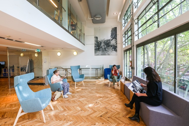 People sitting and reading in a large room with a mezzanine level above and big glass windows to the right.