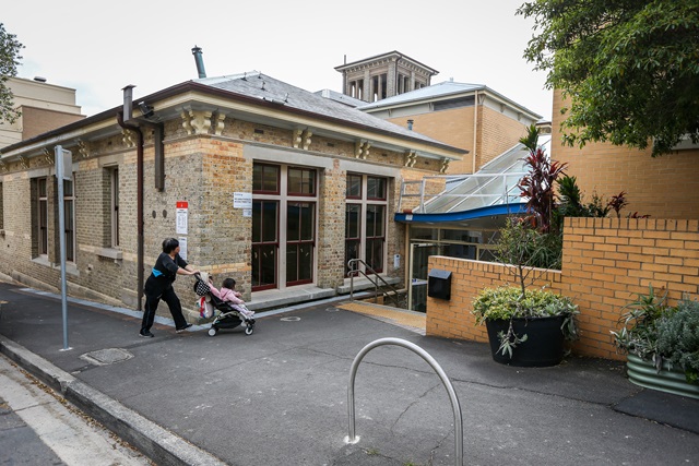 Street view with a footpath, bike rack, buildings and a lady pushing a pram.