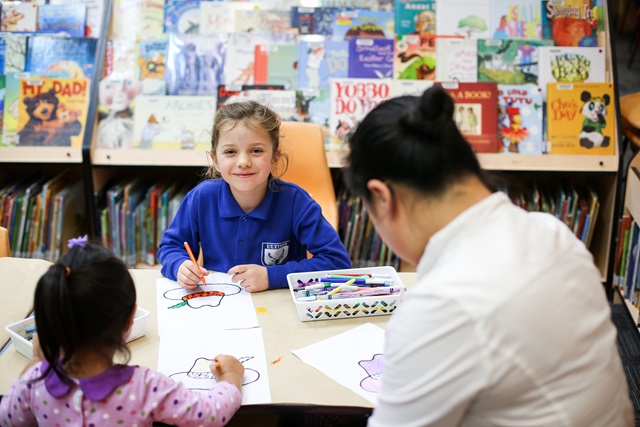 An adult and 2 children one of whom is smiling. Behind her are children's books.