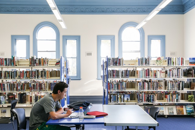 A library area with a man at a desk, behind are shelves of books and large glass windows.