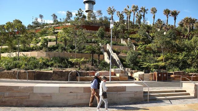 People walking along a path way with a high terraced section in background covered with trees.