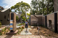 Young woman walking through a paved garden with large trees in the background. 
