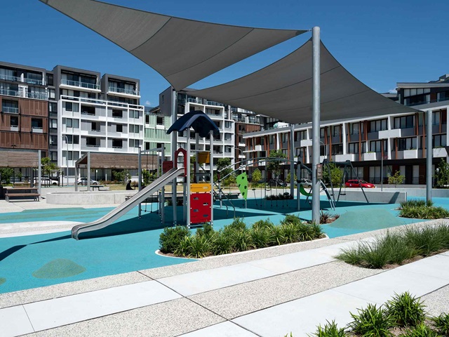 Modern playground with slides and climbing structures under shade sails, surrounded by apartment buildings on a sunny day.