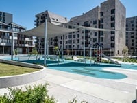 A modern playground with slides and climbing structures is surrounded by apartment buildings. There are shade sails above, and greenery around the area.