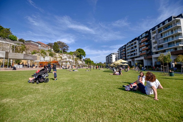 People sit on a grassy lawn on a sunny day. There are small trees and umbrellas for shade. Along the side of the park are apartment buildings.