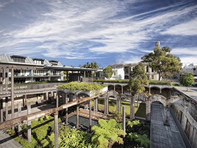 Views from a highpoint looking into a garden integrated into a space made of a mix of old brick archways and industrial metal beams. Busy city road and apartments in the background.