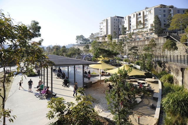 Elevated vantage point with views across a park with families seated under pergolas and seated areas. Apartment blocks in the background.