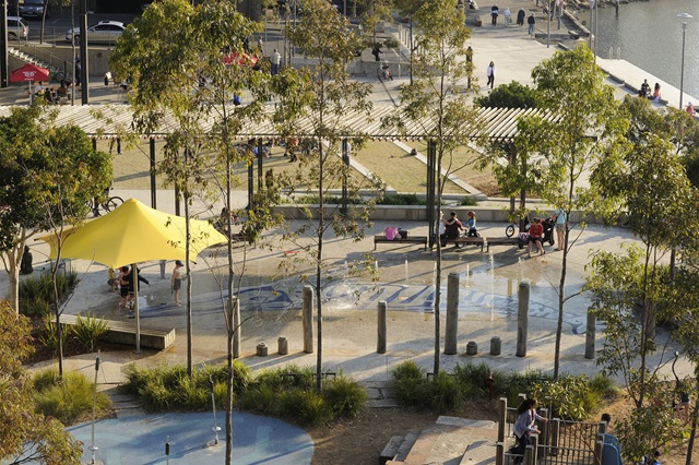 Views over the water feature fountains of the park. tall trees a pergola and seating area surround a walkway of fountains  
