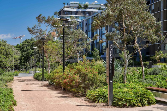A landscaped walking path with modern apartment buildings on the right, greenery on both sides, and a signpost reading "The Rope Walk Park".